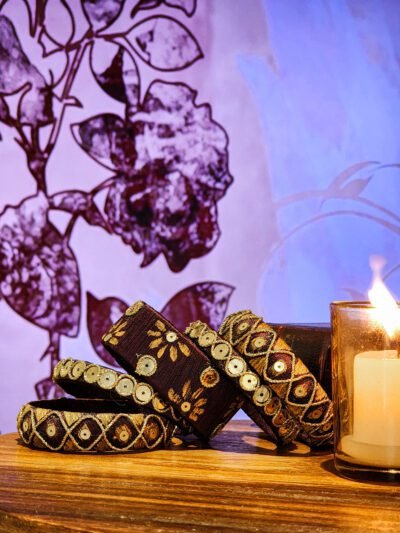 A set of handcrafted maroon velvet bangles adorned with gold sequins, displayed against a soft neutral fabric background. The embroidery reflects South Asian artisan craftsmanship