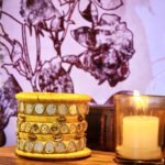 A set of handcrafted gold silk bangles with hand-stitched mirror work and metallic beads, displayed beside a flickering candle on a wooden surface. The intricate patterns reflect South Asian artisan craftsmanship, with soft shadows cast against a backdrop of delicate floral illustrations.
