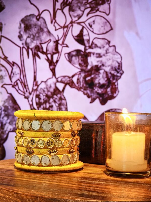 A set of handcrafted gold silk bangles with hand-stitched mirror work and metallic beads, displayed beside a flickering candle on a wooden surface. The intricate patterns reflect South Asian artisan craftsmanship, with soft shadows cast against a backdrop of delicate floral illustrations.