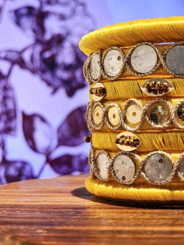 A set of handcrafted gold silk bangles with hand-stitched mirror work and metallic beads, displayed beside a flickering candle on a wooden surface. The intricate patterns reflect South Asian artisan craftsmanship, with soft shadows cast against a backdrop of delicate floral illustrations.