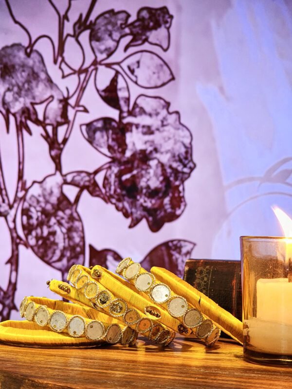 A set of handcrafted gold silk bangles with hand-stitched mirror work and metallic beads, displayed beside a flickering candle on a wooden surface. The intricate patterns reflect South Asian artisan craftsmanship, with soft shadows cast against a backdrop of delicate floral illustrations.