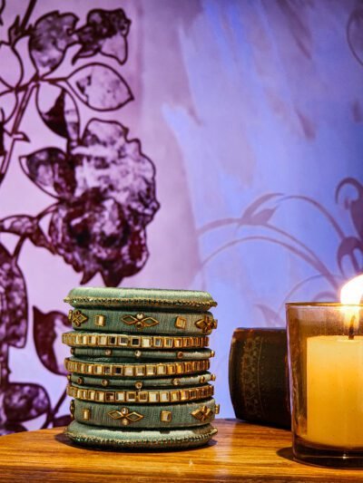 A stack of handwoven periwinkle churas adorned with metallic embroidery and sapphire-toned stones, displayed on a polished wooden surface beside a flickering candle. The intricate patterns and shimmering details highlight masterful artisan craftsmanship