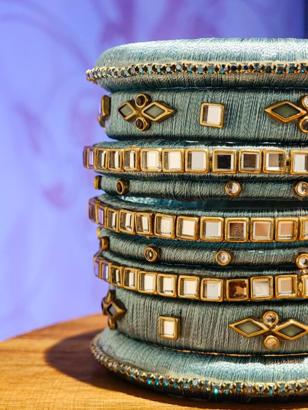 A stack of handwoven periwinkle churas adorned with metallic embroidery and sapphire-toned stones, displayed on a polished wooden surface beside a flickering candle. The intricate patterns and shimmering details highlight masterful artisan craftsmanship