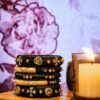 A stack of handcrafted black velvet bangles adorned with gold embellishments and clear stones, displayed on a polished wooden surface beside a flickering candle. The intricate geometric patterns reflect South Asian artisan craftsmanship.