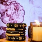A stack of handcrafted black velvet bangles adorned with gold embellishments and clear stones, displayed on a polished wooden surface beside a flickering candle. The intricate geometric patterns reflect South Asian artisan craftsmanship.