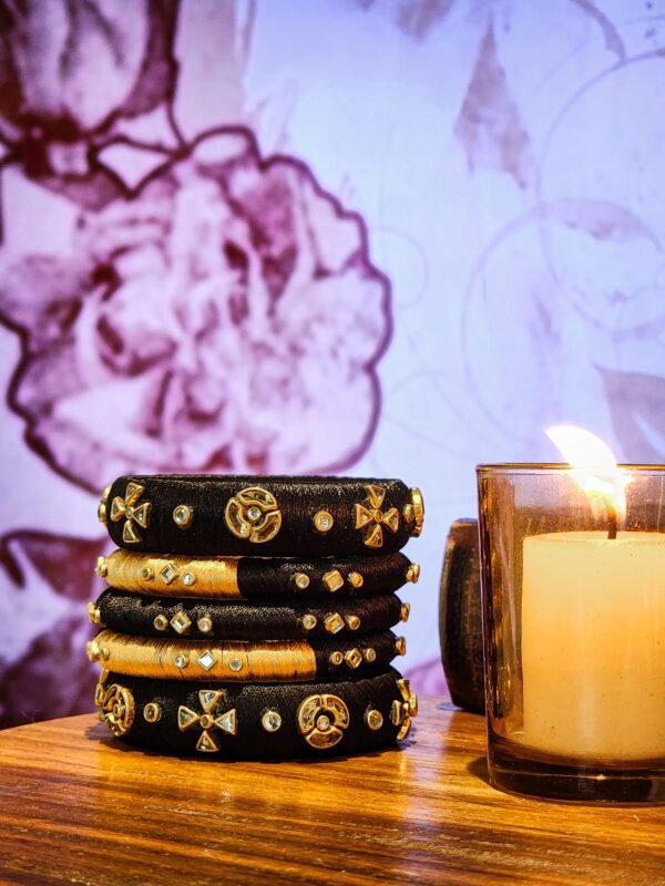 A stack of handcrafted black velvet bangles adorned with gold embellishments and clear stones, displayed on a polished wooden surface beside a flickering candle. The intricate geometric patterns reflect South Asian artisan craftsmanship.