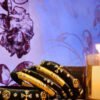 A stack of handcrafted black velvet bangles adorned with gold embellishments and clear stones, displayed on a polished wooden surface beside a flickering candle. The intricate geometric patterns reflect South Asian artisan craftsmanship.