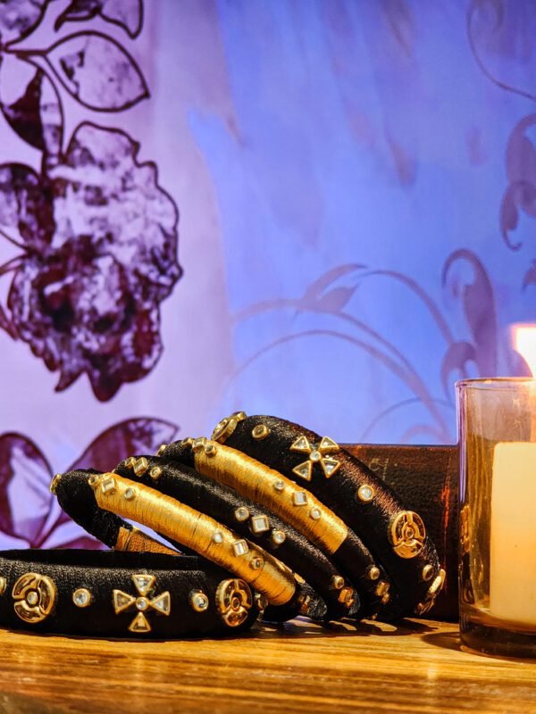A stack of handcrafted black velvet bangles adorned with gold embellishments and clear stones, displayed on a polished wooden surface beside a flickering candle. The intricate geometric patterns reflect South Asian artisan craftsmanship.