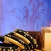 A stack of handcrafted black velvet bangles adorned with gold embellishments and clear stones, displayed on a polished wooden surface beside a flickering candle. The intricate geometric patterns reflect South Asian artisan craftsmanship.