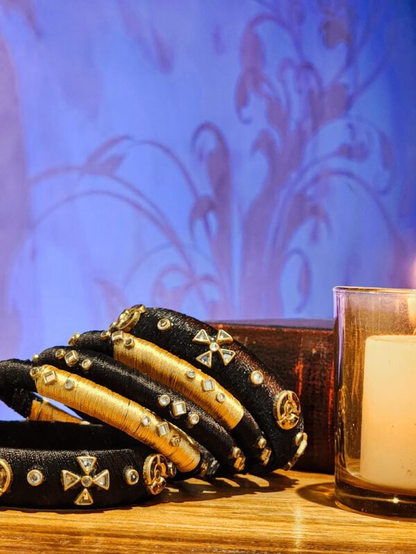 A stack of handcrafted black velvet bangles adorned with gold embellishments and clear stones, displayed on a polished wooden surface beside a flickering candle. The intricate geometric patterns reflect South Asian artisan craftsmanship.