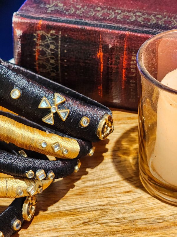 A stack of handcrafted black velvet bangles adorned with gold embellishments and clear stones, displayed on a polished wooden surface beside a flickering candle. The intricate geometric patterns reflect South Asian artisan craftsmanship.