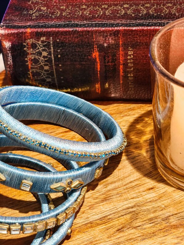 A stack of handwoven periwinkle churas adorned with metallic embroidery and sapphire-toned stones, displayed on a polished wooden surface beside a flickering candle. The intricate patterns and shimmering details highlight masterful artisan craftsmanship