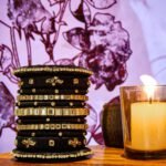 A stack of handcrafted black velvet bangles adorned with gold motifs and crystal accents, displayed on a polished wooden surface beside a flickering candle. The intricate designs reflect the artistry of South Asian craftsmanship.