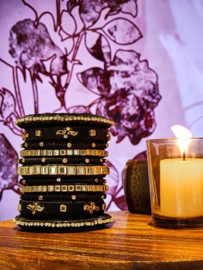 A stack of handcrafted black velvet bangles adorned with gold motifs and crystal accents, displayed on a polished wooden surface beside a flickering candle. The intricate designs reflect the artistry of South Asian craftsmanship.
