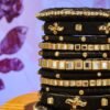 A stack of handcrafted black velvet bangles adorned with gold motifs and crystal accents, displayed on a polished wooden surface beside a flickering candle. The intricate designs reflect the artistry of South Asian craftsmanship.