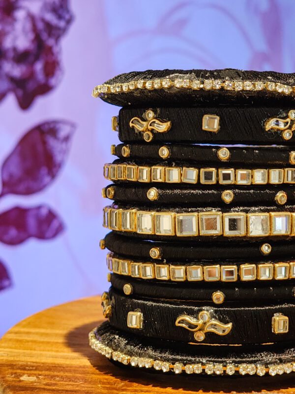 A stack of handcrafted black velvet bangles adorned with gold motifs and crystal accents, displayed on a polished wooden surface beside a flickering candle. The intricate designs reflect the artistry of South Asian craftsmanship.