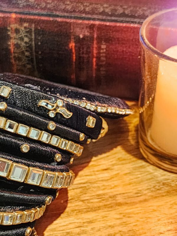 A stack of handcrafted black velvet bangles adorned with gold motifs and crystal accents, displayed on a polished wooden surface beside a flickering candle. The intricate designs reflect the artistry of South Asian craftsmanship.