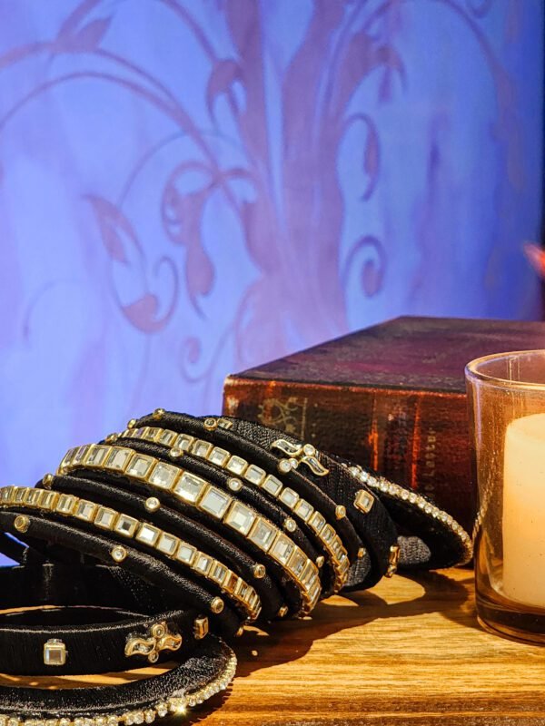 A stack of handcrafted black velvet bangles adorned with gold motifs and crystal accents, displayed on a polished wooden surface beside a flickering candle. The intricate designs reflect the artistry of South Asian craftsmanship.