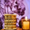A stack of handcrafted beige velvet bangles adorned with hand-embroidered motifs and crystal accents, displayed on a wooden surface beside a flickering candle. The intricate designs reflect traditional South Asian craftsmanship against a backdrop of purple floral patterns.
