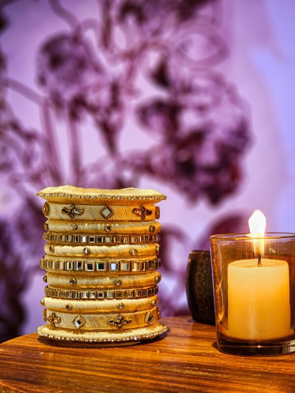 A stack of handcrafted beige velvet bangles adorned with hand-embroidered motifs and crystal accents, displayed on a wooden surface beside a flickering candle. The intricate designs reflect traditional South Asian craftsmanship against a backdrop of purple floral patterns.