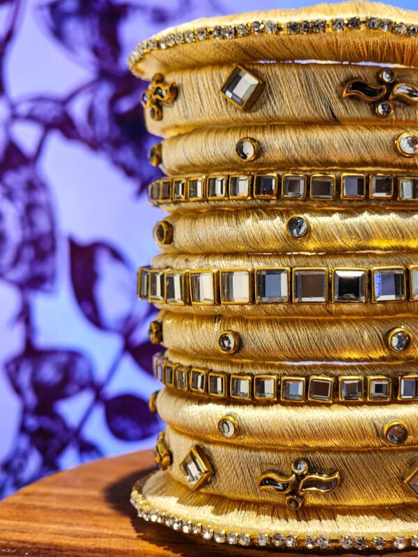 A stack of handcrafted beige velvet bangles adorned with hand-embroidered motifs and crystal accents, displayed on a wooden surface beside a flickering candle. The intricate designs reflect traditional South Asian craftsmanship against a backdrop of purple floral patterns.