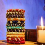 A stack of handcrafted silk bangles in red, gold, blue, and teal, adorned with intricate embroidery and sequins, displayed on a wooden surface beside a flickering candle. The vibrant designs reflect traditional South Asian craftsmanship against a backdrop of soft lighting.