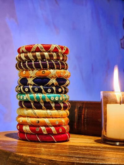 A stack of handcrafted silk bangles in red, gold, blue, and teal, adorned with intricate embroidery and sequins, displayed on a wooden surface beside a flickering candle. The vibrant designs reflect traditional South Asian craftsmanship against a backdrop of soft lighting.
