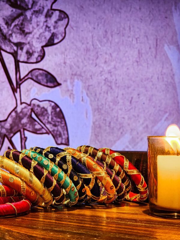 A stack of handcrafted silk bangles in red, gold, blue, and teal, adorned with intricate embroidery and sequins, displayed on a wooden surface beside a flickering candle. The vibrant designs reflect traditional South Asian craftsmanship against a backdrop of soft lighting.