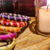 A stack of handcrafted silk bangles in red, gold, blue, and teal, adorned with intricate embroidery and sequins, displayed on a wooden surface beside a flickering candle. The vibrant designs reflect traditional South Asian craftsmanship against a backdrop of soft lighting.