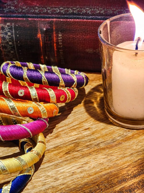 A stack of handcrafted silk bangles in red, gold, blue, and teal, adorned with intricate embroidery and sequins, displayed on a wooden surface beside a flickering candle. The vibrant designs reflect traditional South Asian craftsmanship against a backdrop of soft lighting.