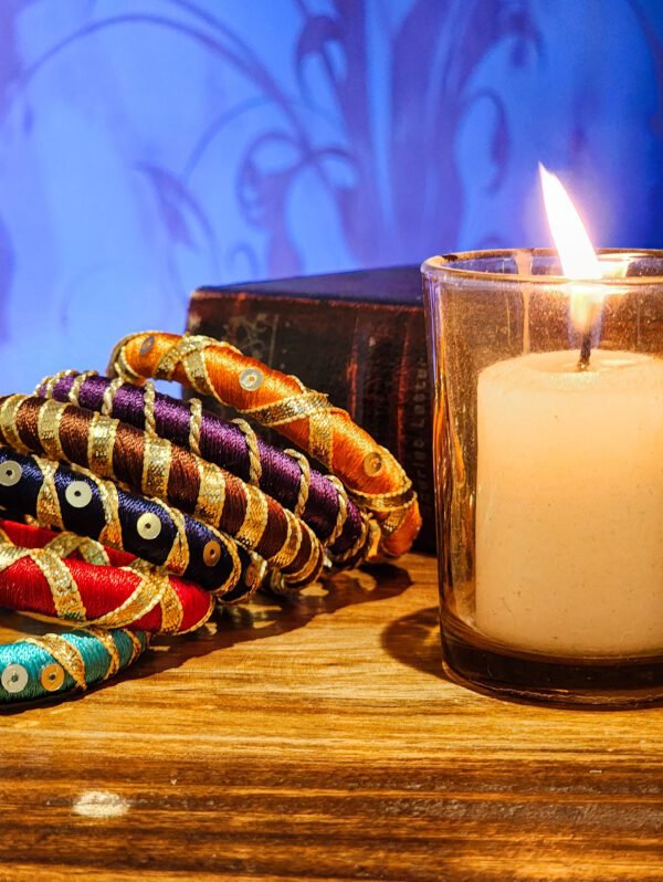 A stack of handcrafted silk bangles in red, gold, blue, and teal, adorned with intricate embroidery and sequins, displayed on a wooden surface beside a flickering candle. The vibrant designs reflect traditional South Asian craftsmanship against a backdrop of soft lighting.