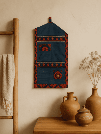 Handmade denim wall hanging with traditional Ajrak fabric detailing, featuring multiple pockets, hanging on a textured beige wall next to wooden and terracotta home decor elements — a cozy, artisan-inspired interior by Jharoka Studio.