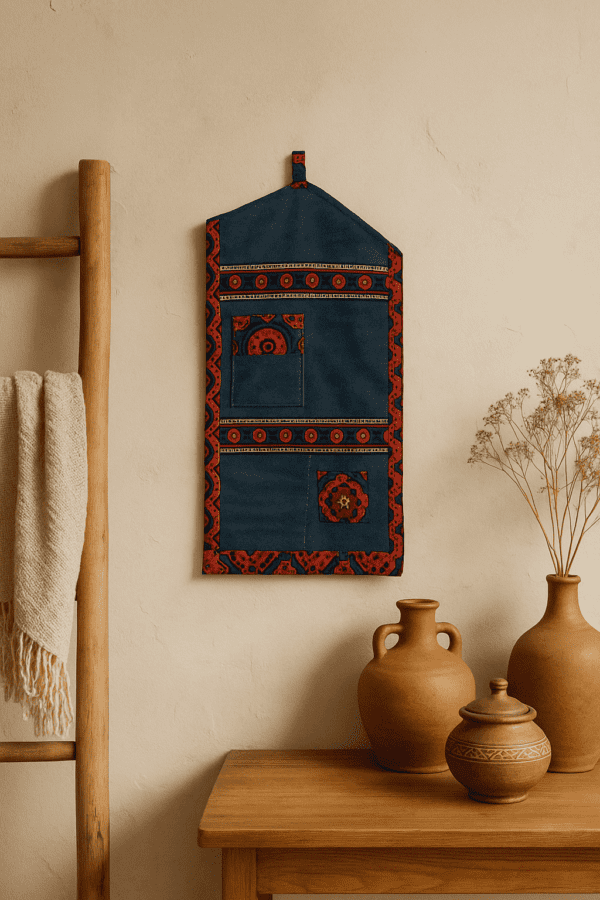 Handmade denim wall hanging with traditional Ajrak fabric detailing, featuring multiple pockets, hanging on a textured beige wall next to wooden and terracotta home decor elements — a cozy, artisan-inspired interior by Jharoka Studio.
