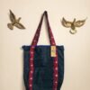 A denim tote bag with Ajrak patterned strips. with Jharoka Studio tag attached framed between two golden birs in a studio wall