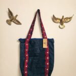 A denim tote bag with Ajrak patterned strips. with Jharoka Studio tag attached framed between two golden birs in a studio wall
