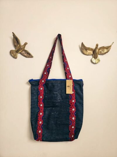 A denim tote bag with Ajrak patterned strips. with Jharoka Studio tag attached framed between two golden birs in a studio wall