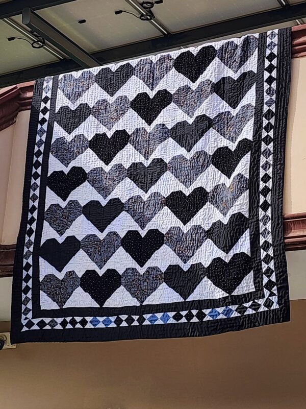 A handcrafted tapestry quilt featuring bold black and white heart patterns, adorned with intricate embroidery details. Displayed against a rustic backdrop, showcasing its versatility as both bedding and wall art.