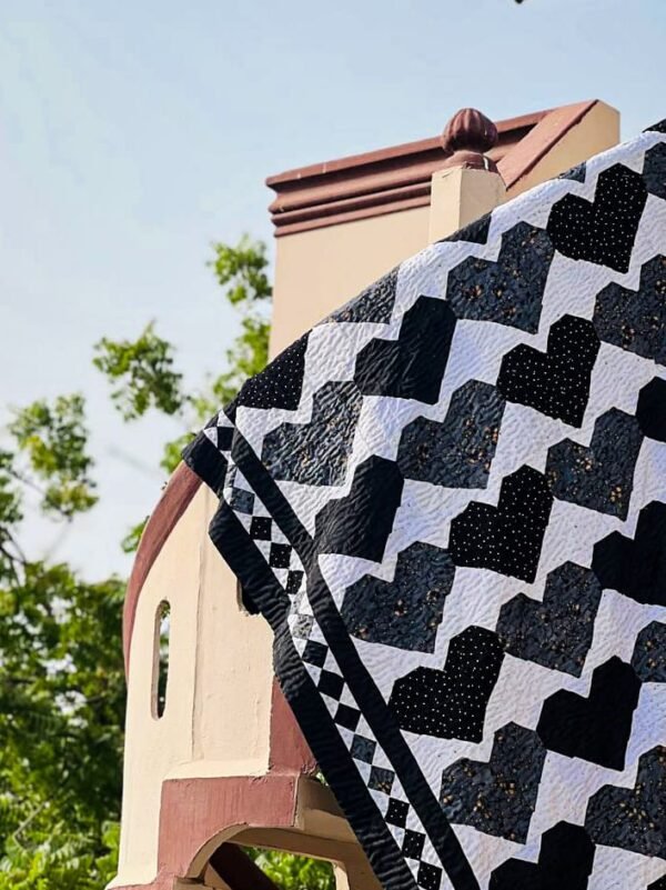 A handcrafted tapestry quilt featuring bold black and white heart patterns, adorned with intricate embroidery details. Displayed against a rustic backdrop, showcasing its versatility as both bedding and wall art.