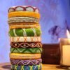 A stack of handcrafted thread bangles in green, orange, blue, and purple, adorned with intricate embroidery and sequins, displayed on a wooden surface beside a flickering candle. The vibrant designs reflect traditional South Asian craftsmanship against a backdrop of soft lighting.