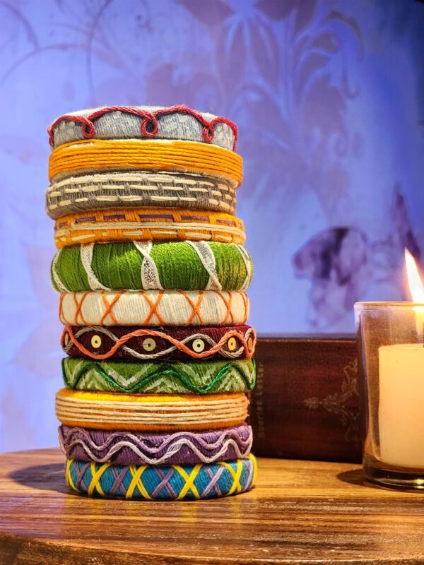 A stack of handcrafted thread bangles in green, orange, blue, and purple, adorned with intricate embroidery and sequins, displayed on a wooden surface beside a flickering candle. The vibrant designs reflect traditional South Asian craftsmanship against a backdrop of soft lighting.