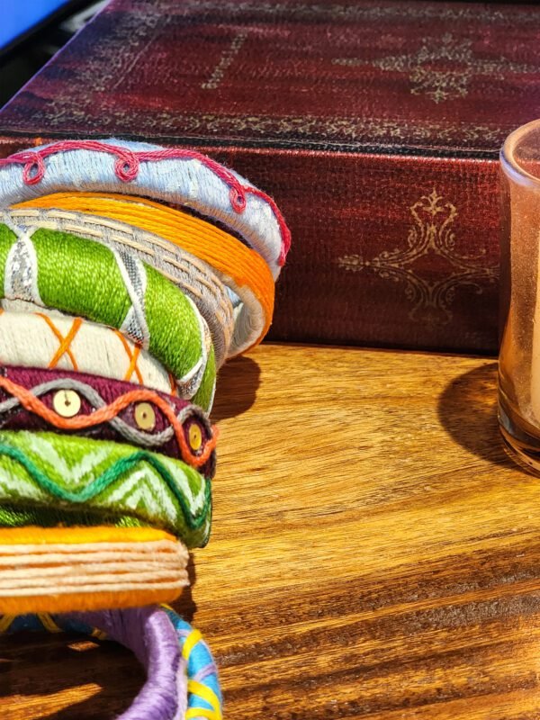 A stack of handcrafted thread bangles in green, orange, blue, and purple, adorned with intricate embroidery and sequins, displayed on a wooden surface beside a flickering candle. The vibrant designs reflect traditional South Asian craftsmanship against a backdrop of soft lighting.