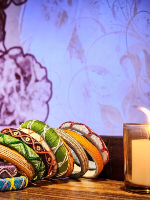 A stack of handcrafted thread bangles in green, orange, blue, and purple, adorned with intricate embroidery and sequins, displayed on a wooden surface beside a flickering candle. The vibrant designs reflect traditional South Asian craftsmanship against a backdrop of soft lighting.