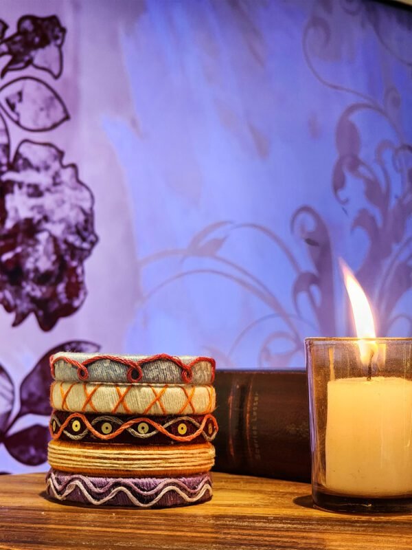 A stack of handcrafted thread bangles in green, orange, blue, and purple, adorned with intricate embroidery and sequins, displayed on a wooden surface beside a flickering candle. The vibrant designs reflect traditional South Asian craftsmanship against a backdrop of soft lighting.