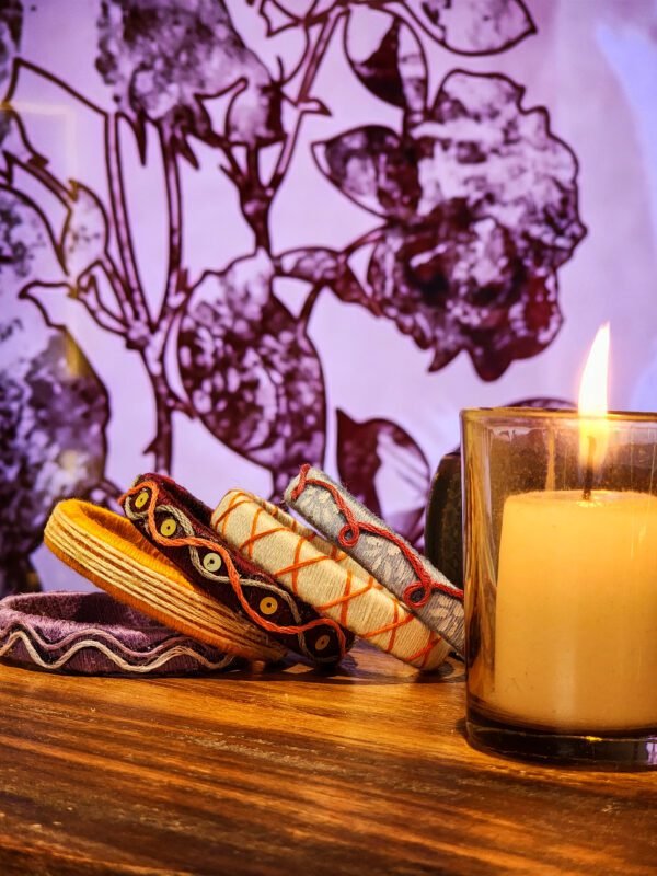 A stack of handcrafted thread bangles in green, orange, blue, and purple, adorned with intricate embroidery and sequins, displayed on a wooden surface beside a flickering candle. The vibrant designs reflect traditional South Asian craftsmanship against a backdrop of soft lighting.