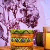 A stack of handcrafted thread bangles in green, orange, blue, and purple, adorned with intricate embroidery and sequins, displayed on a wooden surface beside a flickering candle. The vibrant designs reflect traditional South Asian craftsmanship against a backdrop of soft lighting.
