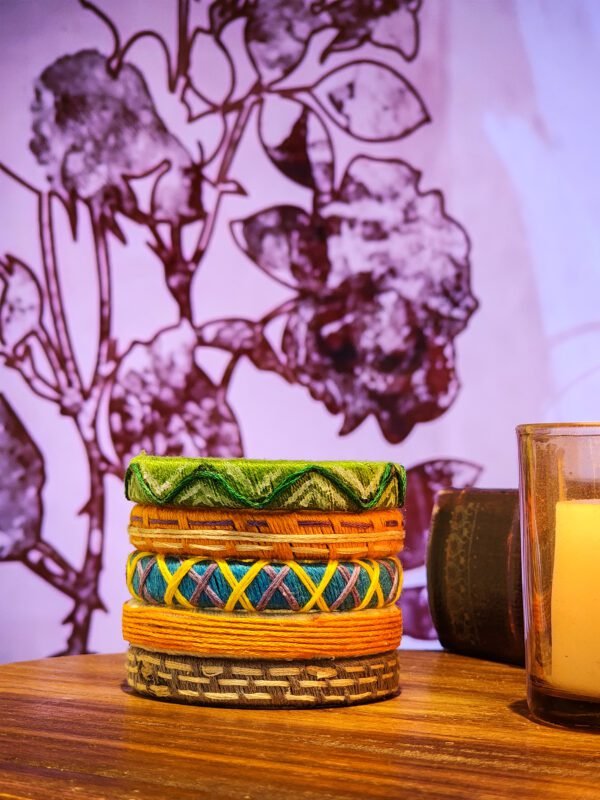 A stack of handcrafted thread bangles in green, orange, blue, and purple, adorned with intricate embroidery and sequins, displayed on a wooden surface beside a flickering candle. The vibrant designs reflect traditional South Asian craftsmanship against a backdrop of soft lighting.