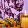 A stack of handcrafted thread bangles in green, orange, blue, and purple, adorned with intricate embroidery and sequins, displayed on a wooden surface beside a flickering candle. The vibrant designs reflect traditional South Asian craftsmanship against a backdrop of soft lighting.