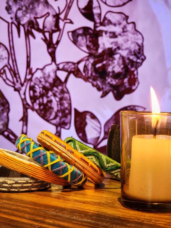 A stack of handcrafted thread bangles in green, orange, blue, and purple, adorned with intricate embroidery and sequins, displayed on a wooden surface beside a flickering candle. The vibrant designs reflect traditional South Asian craftsmanship against a backdrop of soft lighting.