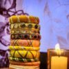 A stack of handcrafted thread bangles in gold, red, blue, and green, adorned with mirrorwork, sequins, and intricate embroidery, displayed on a wooden surface beside a flickering candle. The warm glow highlights the artisanal craftsmanship of South Asian textiles.