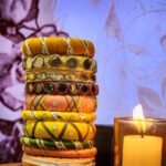 A stack of handcrafted thread bangles in gold, red, blue, and green, adorned with mirrorwork, sequins, and intricate embroidery, displayed on a wooden surface beside a flickering candle. The warm glow highlights the artisanal craftsmanship of South Asian textiles.
